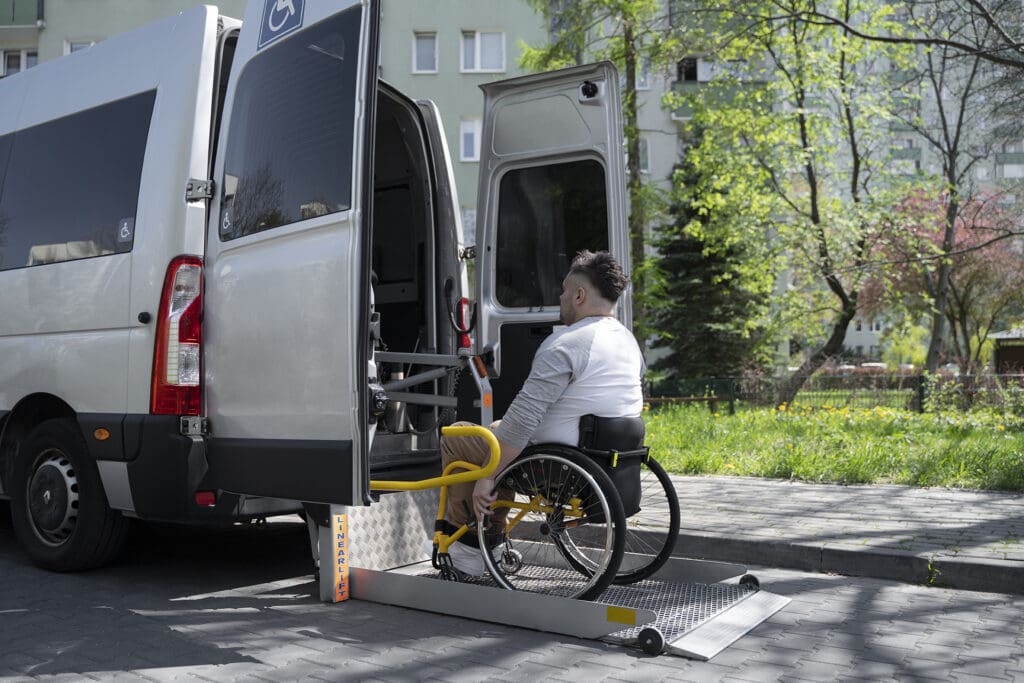 A man in a wheelchair ascends a van ramp while a staff member stands nearby.