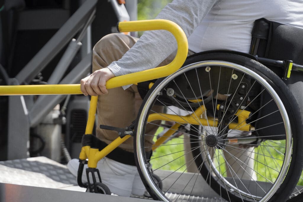 A man in a wheelchair ascends a van ramp while a staff member stands nearby.